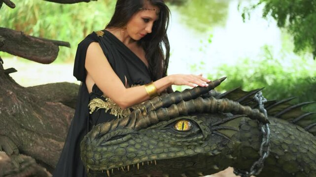Portrait art Fantasy woman queen hand stroking touching her dragon head muzzle pet animal with love tamed dragon slave neck in collar sits on chain. summer nature forest green trees. Black dress 4k