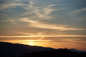 Sun sets behind rolling hills, casting warm, golden glow across sky. Wispy clouds, tinged with hues of orange and yellow, stretch across deepening blue sky. Serene and expansive landscape.