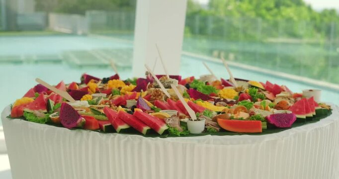 Medium shot of a table with snacks in a wedding banquet hall against the background of a window with a pool view. Offsite catering for celebrations. Meat, tropical fruits and canapes on the table.