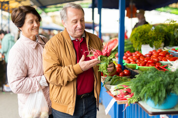 Elderly married couple in casual clothes choosing organic radish near vegetable stand at local grocery market