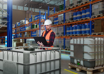 Man in chemical warehouse. Storekeeper with laptop stands among barrels. Fuel warehouse inside IBC. Man warehouse manager at work. Guy inside storage hangar. Supervisor among racks of barrels