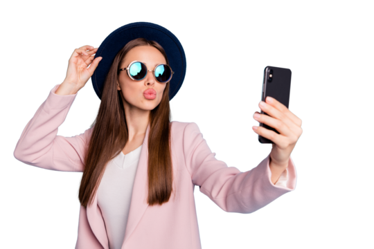 Portrait of dreamy cute girl using her mobile phone making selfie sending air kisses launching blog stream wearing pink stylish topcoat isolated over blue background