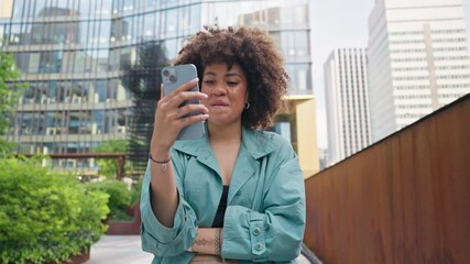 Satisfied African American woman enjoying using phone during walk on street. Pretty girl holding smartphone while watching podcast or video on Internet. With positive smile spending time outside.