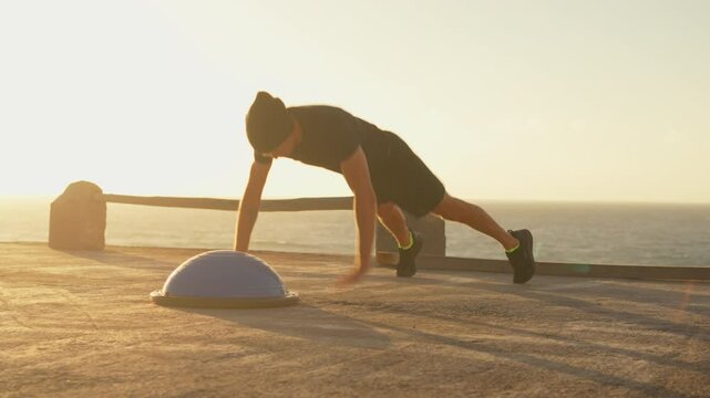 Determined athlete in sportswear doing pushups using half ball balance trainer. Sporty young man exercising near sea under sky during sunset. Fitness and healthy lifestyle.