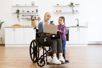 Woman with disability in a wheelchair using laptop in kitchen with young girl. Warm family interaction and technology use. Bright and modern kitchen setting.