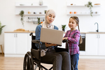 Woman in wheelchair working on laptop with young girl assisting in modern kitchen. Concept of...