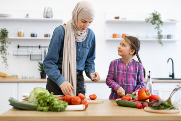 Smiling daughter helps mother with cooking. Muslim mother and daughter cutting fresh ingredients for salad in kitchen. Family bonding over healthy meal preparation, promoting healthy eating habits.