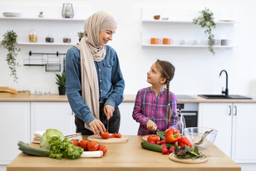 Fresh vegetables on table, smiling and working together creating healthy meal, enjoying family time in modern kitchen. Muslim mother and daughter cutting ingredients for salad.