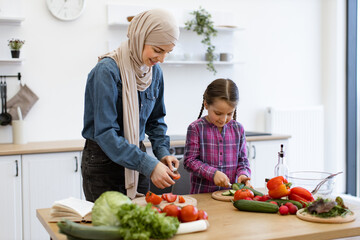 Muslim mother and daughter cutting ingredients for salad in bright modern kitchen. Both are smiling and enjoying quality time together while preparing healthy meal.