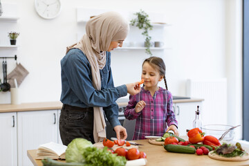 Family bonding while preparing fresh vegetables. Concept of healthy eating, family time, and cultural traditions. Muslim mother and daughter cutting ingredients for salad in modern kitchen.