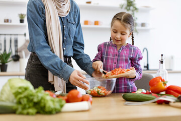 Family bonding moment in bright kitchen setting promoting healthy eating and cooking together. Muslim mother and daughter pour chopped vegetables from wooden board into salad bowl.