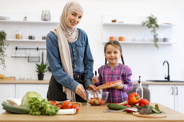 Smiling family bonding while cooking healthy meal. Muslim mother and daughter pour chopped vegetables from wooden board into salad bowl. Concept of healthy eating, family time, and cultural traditions