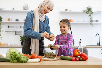 Muslim mother and daughter chop, tear greens in salad bowl. They enjoy family time together while...