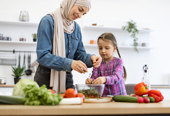 Fresh vegetables on kitchen counter. Muslim mother and daughter chop, tear greens in salad bowl. Bonding activity promotes healthy eating, family values.