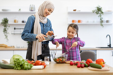 Muslim mother and daughter add olive oil to salad bowl. Family bonding while preparing fresh vegetable salad. Teaching cooking skills and healthy eating habits.