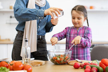 Muslim mother and daughter adding salt to salad bowl in kitchen. Mother wearing hijab and daughter smiling while stirring fresh vegetable salad. Concept of family bonding, healthy eating at home.