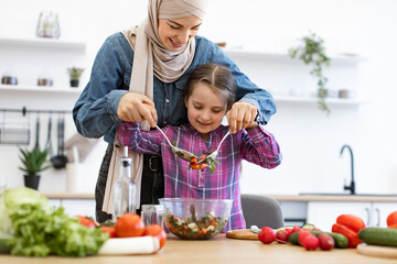 Image depicts cooking, togetherness, healthy eating. Muslim mother and daughter mixing chopped vegetables in salad bowls in kitchen.Family bonding and joy in cooking at home.