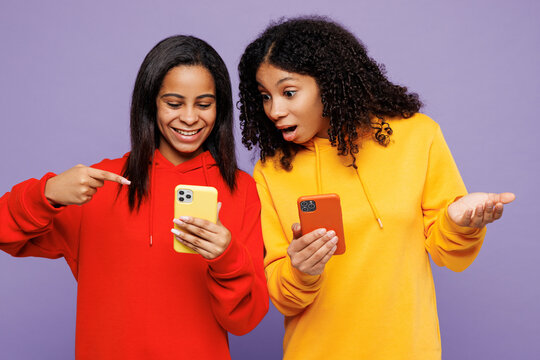 Two young amazed little kid teen girls women they wear red yellow hoodie casual clothes hold in hand use point on mobile cell phone isolated on plain pastel light purple background. Lifestyle concept.