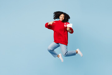 Full body little kid teen girl of African American ethnicity wear red sweatshirt jump high hold point gift coupon voucher card for store isolated on plain blue background. Childhood lifestyle concept.