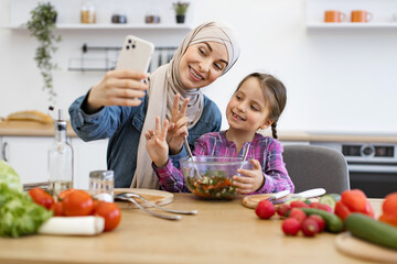 Smiling faces, bonding moment, healthy lifestyle, and family connection highlighted in bright. Muslim mother and daughter engaging in video call showcasing freshly prepared healthy salad in kitchen.