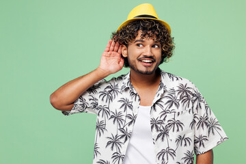 Young curious nosy fun Indian man wear beach shirt hat rest near hotel pool try to hear you overhear listening intently isolated on plain green color background. Summer vacation sea sun tan concept.