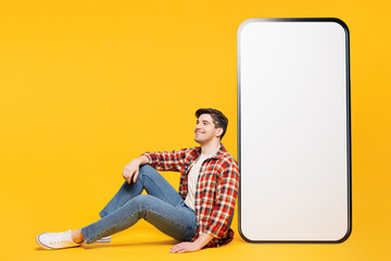 Full body side view young man he wears red checkered shirt casual clothes sit near big huge blank screen mobile cell phone smartphone with area isolated on plain yellow background. Lifestyle concept.