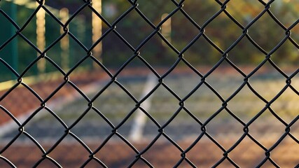 Chain-link fence with tennis court in the background, sunlit evening. Urban sport and recreation concept