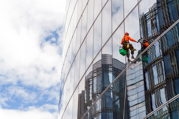 Worker in safety gear clean the windows of a tall glass building, suspended high above the ground.