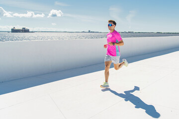 A man in a bright pink shirt jogs along a sunlit waterfront, exuding energy and determination.