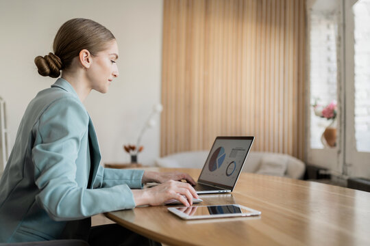 A professional woman in a light blue suit works on a laptop, analyzing data charts in a modern, minimalistic office.