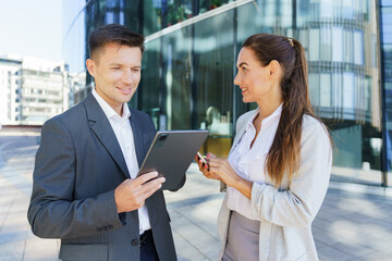 Two business professionals, a man and a woman, engaging in a discussion outdoors while using a tablet and smartphone, set against a modern glass building.