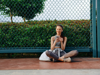 Young woman sitting on the ground, smiling while using her smartphone, surrounded by greenery and a...