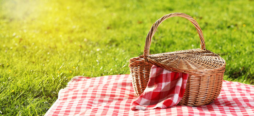 Picnic blanket and wicker basket on green grass during sunny day. Banner design with space for text