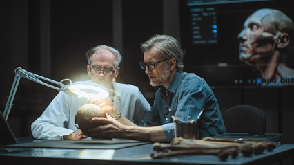 Two archaeologists take measurements of ancient human skull, study fossil remains, use tablet...
