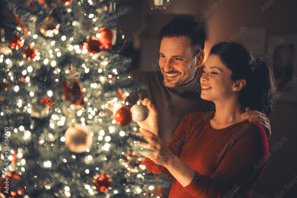 Wall mural A lovely happy young adult married couple decorating the Christmas tree in their cozy home