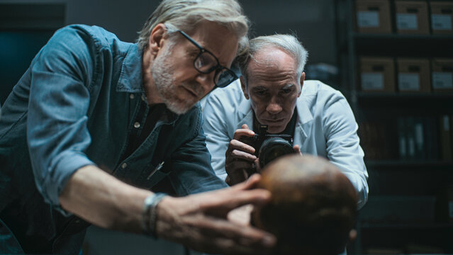 Modern archaeological laboratory: Two archaeologists set up professional camera to photograph fossil remains, have discussion. Senior scientist in lab coat takes photo with flash. Archaeology concept. - Powered by Adobe