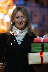 Happy smiling young blonde woman with gift boxes and a sparkler near the Christmas tree at night in the city. Concept of holidays, Christmas, shopping. Vertical photo