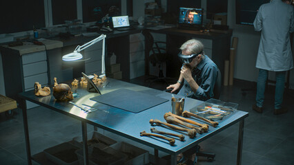 Archaeological lab at night: Mature archaeologist or scientist in AR headset conducts historical...