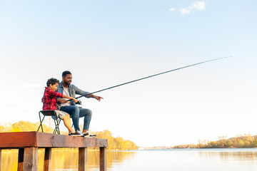 An African American father and his son sit on a wooden pier by a lake. The father, wearing a denim...