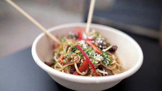 A delicious bowl of Asian noodles topped with fresh vegetables and sesame seeds, served with chopsticks in a biodegradable container. High quality 4k footage