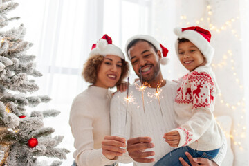 african american family in santa hats celebrating new year together and holding sparklers at home near christmas tree, mom dad and little son having fun at christmas