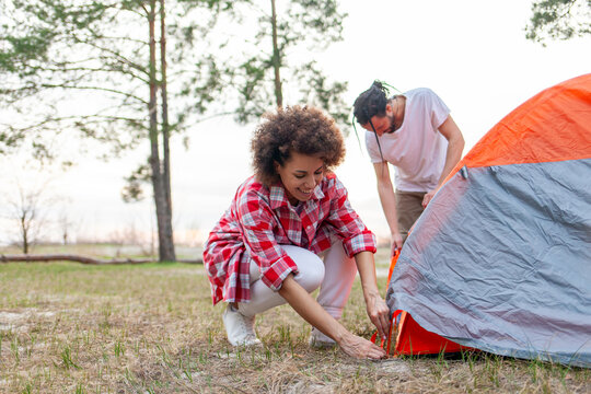 young african american couple hikers pitching tent in forest near lake, man and woman with camping equipment and backpacks traveling and preparing camping site - Powered by Adobe