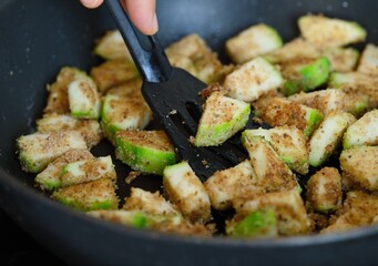 A woman hand with a spatula stirring breaded pieces of courgette that are being fried in a frying pan. Close-up