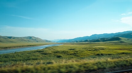 Fototapeta premium A scenic view from a train window, capturing rolling green hills, distant mountains, and a river running parallel to the tracks, under a clear blue sky.