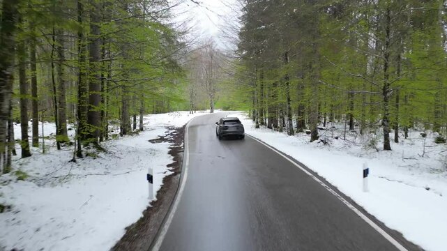 suv driving on road in snow surrounded by fores trees low over the street chasing following aerial shot winter landscape