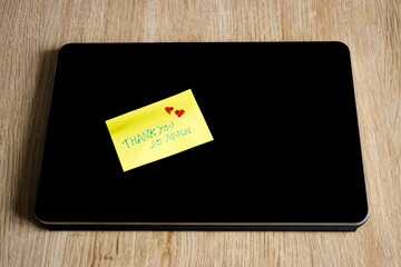 Vista superior de una nota adhesiva amarilla con agradecimiento y corazones sobre un dispositivo tablet con fondo de madera