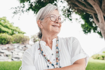 Portrait of attractive smiling senior woman of 70 years old sitting in the park looking away. Elderly lady enjoying a sunny day in outdoors