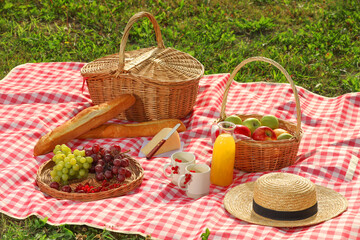 Picnic basket, different snacks, juice and straw hat on red checkered blanket outdoors