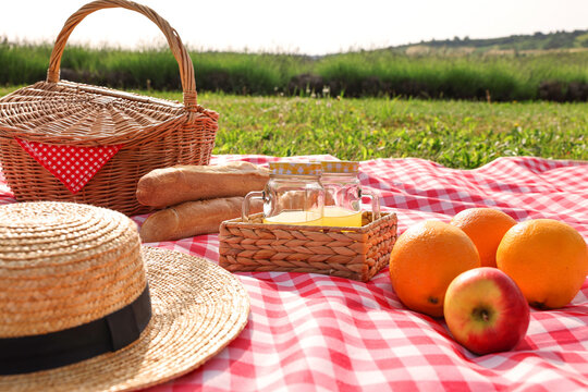Picnic basket, different snacks, juice and straw hat on red checkered blanket outdoors