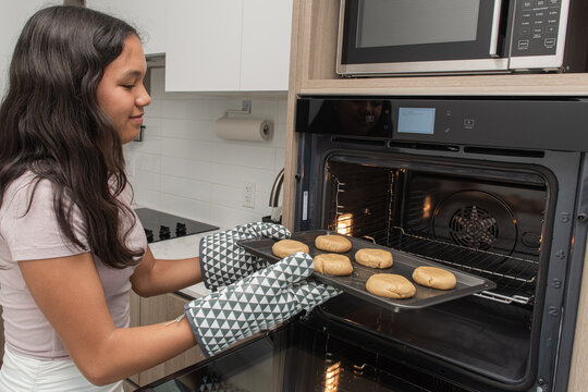 Young teenager wearing mittens putting raw cookies on the hot oven.